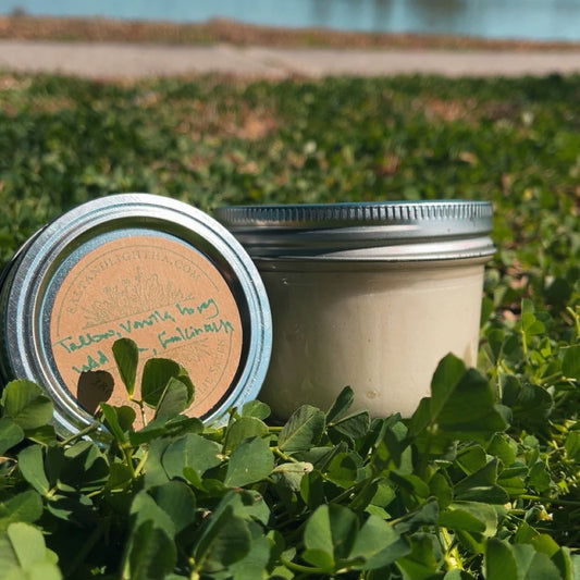 Two metal jars of Honey Vanilla Tallow lotion on a bed of green leaves with a lake and trees in the background