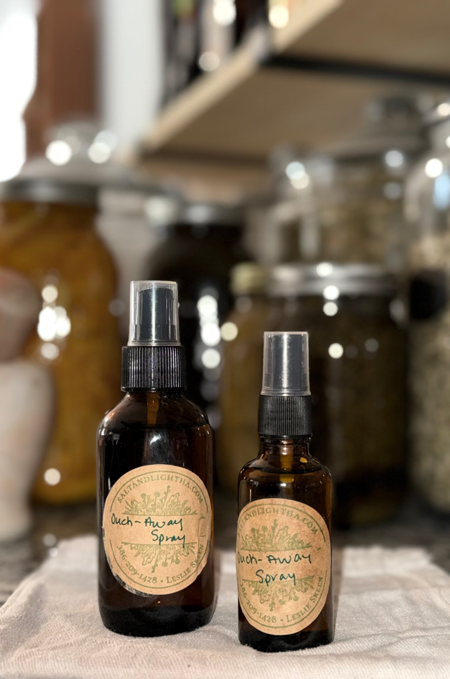 Two brown spray bottles with labels that say ouch-away spray with jars in the background