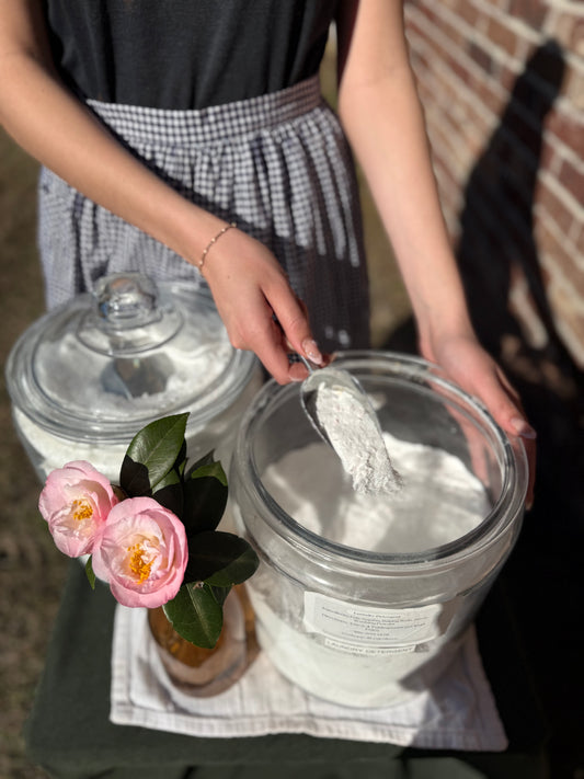 Person holding two large glass jars of powdered laundry soap with floral decorations and a checkered apron. At the Salt and Light House