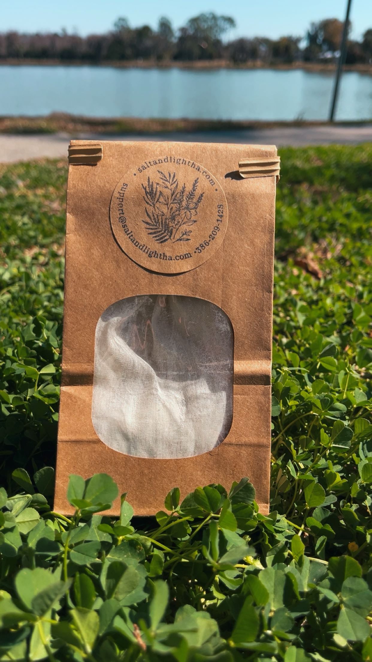  paper packaging with a window showing a Bath Salt, placed on grass with a lake in the background.