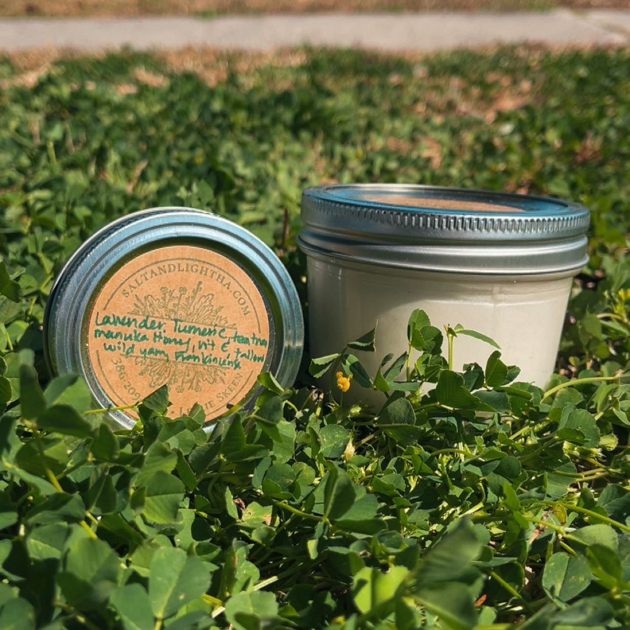 Two jars of Lavender Turmeric on a bed of green plants with a body of water and trees in the background.