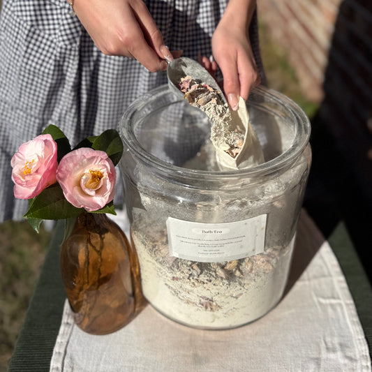 Person scooping a bath tea from a jar with flowers and a bottle nearby.